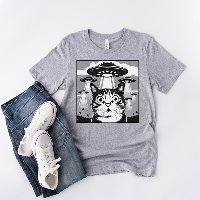 Puede incluir: Camiseta gris claro con un dise&ntilde;o gr&aacute;fico en blanco y negro de un gato sorprendido mirando varios ovnis. El gato tiene ojos grandes. La camiseta se combina con vaqueros azules y zapatillas blancas.
