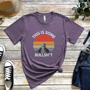 May include: Purple t-shirt with a retro sunset graphic and the text "This is some bullsh*t". The graphic features a blue alien character.