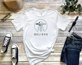 May include: White t-shirt with a black and white illustration of a UFO with the word "BELIEVE" printed below it. The t-shirt is laid flat on a wooden surface with a pair of black sneakers, a straw hat, a pair of glasses, a watch, and a pair of earbuds.