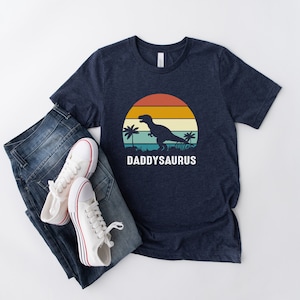 Puede incluir: Una camiseta azul marino con un gráfico de puesta de sol retro que presenta una silueta de dinosaurio y el texto "DADDYSAURUS".