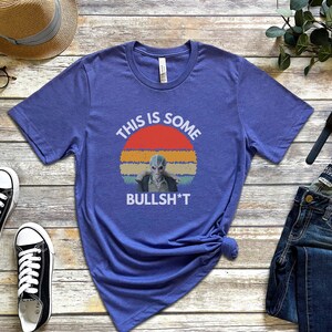 May include: A blue t-shirt with a retro sunset graphic and the text "This is some bullsh*t". The graphic features a blue alien with a serious expression.