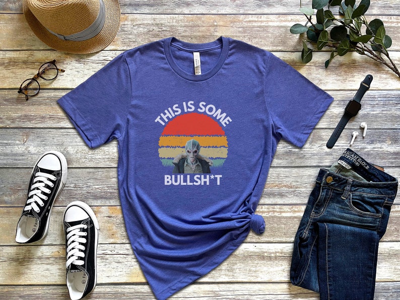 May include: A blue t-shirt with a retro sunset graphic and the text "This is some bullsh*t". The graphic features a blue alien with a serious expression.