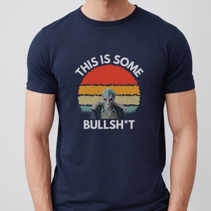 May include: A navy blue t-shirt with a retro sunset graphic and the text "This is some BULLSH*T". The graphic features a blue alien character.