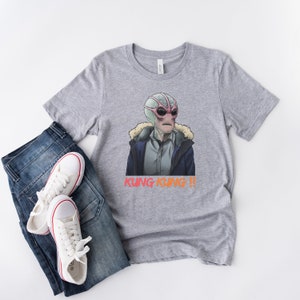 May include: A grey t-shirt with a graphic of a pink and green alien wearing a blue jacket and the text "Kung Kung!!" printed in orange and red.