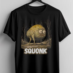May include: Black t-shirt with a cartoon illustration of a green, lumpy creature with large eyes. The creature is walking on a dirt path in a forest. The text "SQUONK" is printed below the creature.