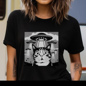 Puede incluir: Camiseta negra con un gr&aacute;fico en blanco y negro de un gato mirando platillos volantes. El gato tiene ojos grandes y el dise&ntilde;o incluye rayos de luz de los ovnis. El fondo muestra un cielo nublado.