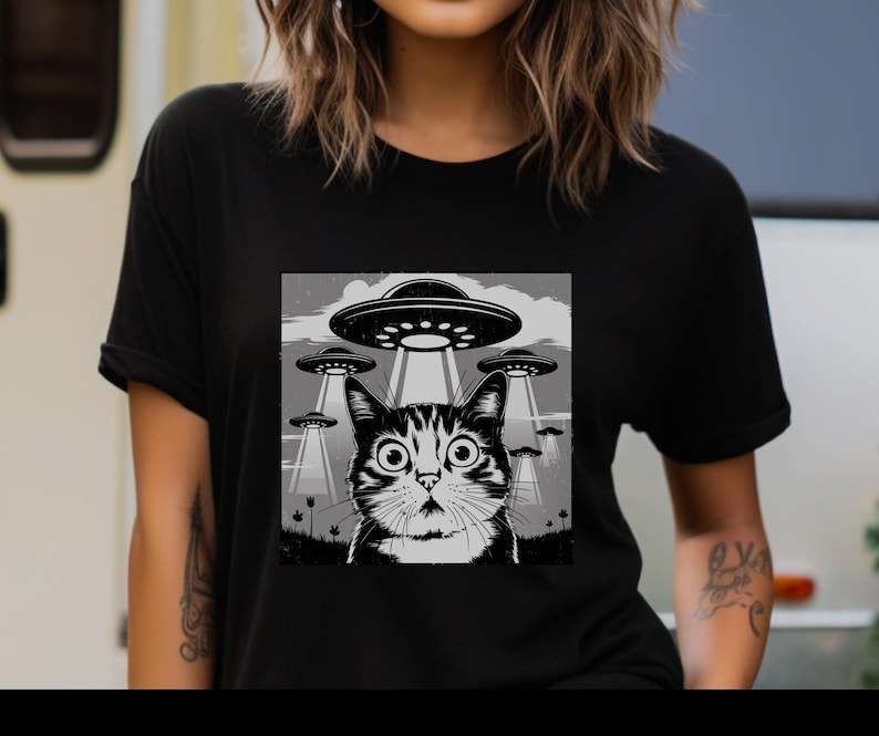 Puede incluir: Camiseta negra con un gr&aacute;fico en blanco y negro de un gato mirando platillos volantes. El gato tiene ojos grandes y el dise&ntilde;o incluye rayos de luz de los ovnis. El fondo muestra un cielo nublado.