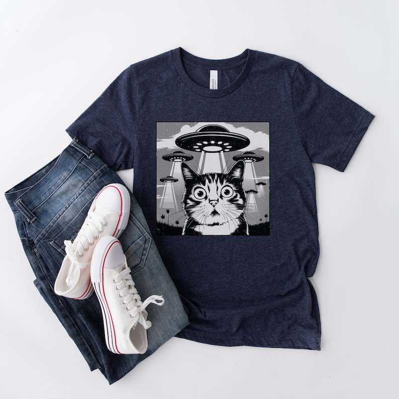 Puede incluir: Camiseta azul marino con un gr&aacute;fico en blanco y negro de un gato sorprendido y OVNIs. La camiseta se muestra con vaqueros azules y zapatillas blancas con detalles rojos. La camiseta est&aacute; hecha de un material suave.