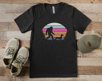 May include: A dark grey t-shirt featuring a retro sunset graphic with a silhouette of Bigfoot walking a dachshund.