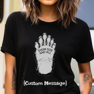 May include: Black t-shirt with a graphic of a large, furry, gray foot print. The text "Show Size And All" is printed on the foot print. The text "{Custom Message}" is printed below the graphic.