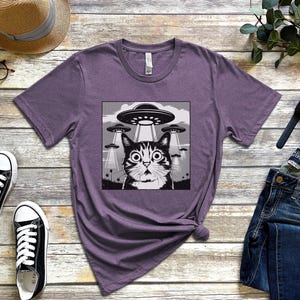 Puede incluir: Camiseta morada con un dise&ntilde;o gr&aacute;fico en blanco y negro de un gato sorprendido y OVNIs. La camiseta est&aacute; hecha de material suave. La imagen tambi&eacute;n muestra un par de zapatillas negras, un sombrero de paja y vaqueros azules. Adecuado para uso diario.