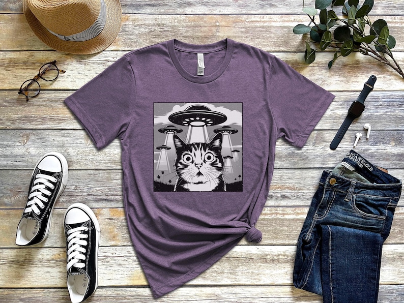 Puede incluir: Camiseta morada con un dise&ntilde;o gr&aacute;fico en blanco y negro de un gato sorprendido y OVNIs. La camiseta est&aacute; hecha de material suave. La imagen tambi&eacute;n muestra un par de zapatillas negras, un sombrero de paja y vaqueros azules. Adecuado para uso diario.