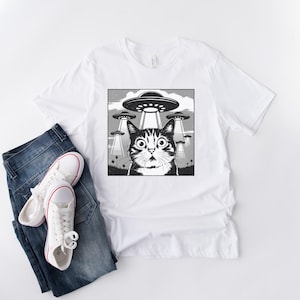 Puede incluir: Camiseta blanca con un gr&aacute;fico en blanco y negro de un gato sorprendido mirando ovnis. El gato tiene ojos grandes y est&aacute; centrado en un dise&ntilde;o cuadrado. La camiseta se combina con vaqueros azules y zapatillas blancas.