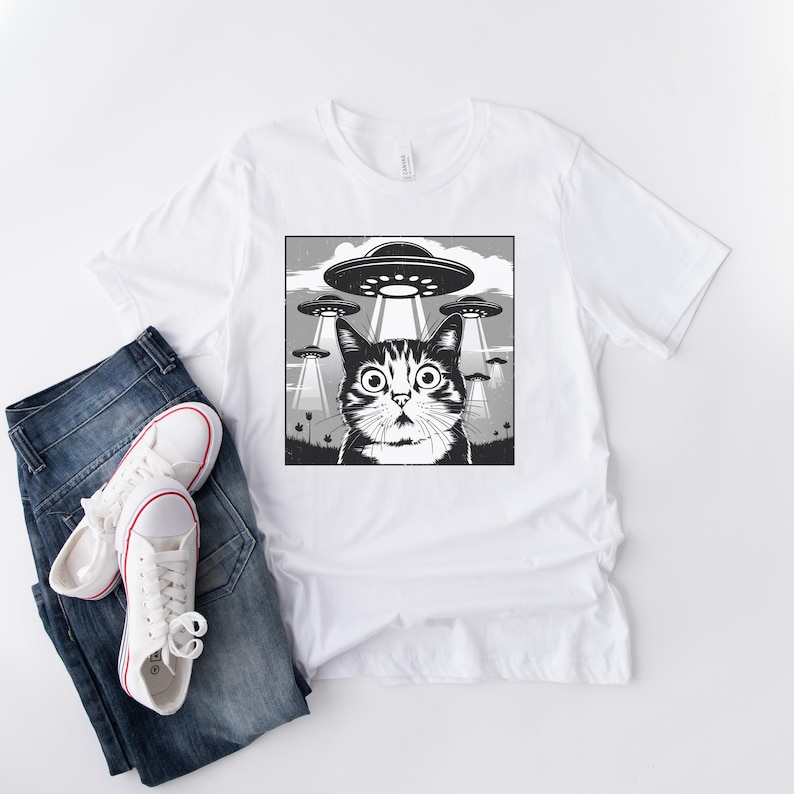Puede incluir: Camiseta blanca con un gr&aacute;fico en blanco y negro de un gato sorprendido mirando ovnis. El gato tiene ojos grandes y est&aacute; centrado en un dise&ntilde;o cuadrado. La camiseta se combina con vaqueros azules y zapatillas blancas.