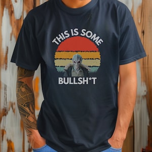 May include: A navy blue t-shirt with a vintage-style sunset graphic and the text "This is some BULLSH*T". The graphic features a gray alien figure in the center.