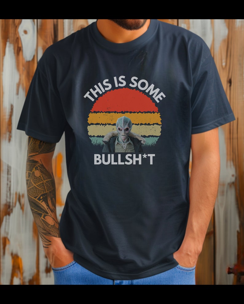 May include: A navy blue t-shirt with a vintage-style sunset graphic and the text "This is some BULLSH*T". The graphic features a gray alien figure in the center.
