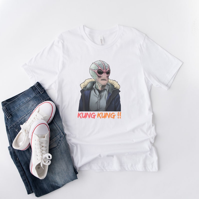 May include: White t-shirt with a cartoon illustration of a creature with a pink and green head and large eyes. The creature is wearing a blue jacket and the text "KUNG KUNG!!" is printed below the image.