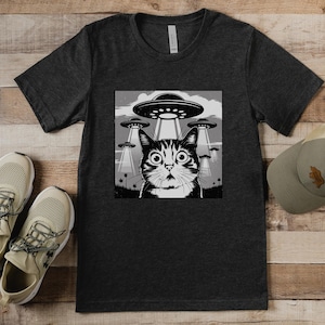 Puede incluir: Camiseta gris oscuro con un gr&aacute;fico en blanco y negro de un gato sorprendido mirando ovnis. Una gorra y zapatillas a juego est&aacute;n al lado de la camiseta. La camiseta est&aacute; hecha de un material suave. Las zapatillas son beige.