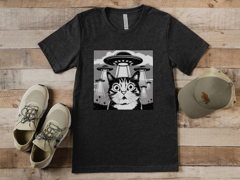 Puede incluir: Camiseta gris oscuro con un gr&aacute;fico en blanco y negro de un gato sorprendido mirando ovnis. Una gorra y zapatillas a juego est&aacute;n al lado de la camiseta. La camiseta est&aacute; hecha de un material suave. Las zapatillas son beige.