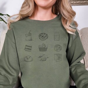 May include: A green sweatshirt with a graphic print of various food items, including a loaf of bread, a basket of bread, a jar of jam, and a pastry.