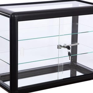 May include: A black display case with glass panels and shelves. The case has a lockable door with a key. The frame is black, and the shelves are clear glass. Ideal for displaying collectibles or merchandise.