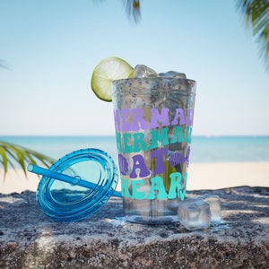 Op de afbeelding: Een doorzichtige plastic beker met een blauwe deksel en rietje. Op de beker staat "Mermaid at Heart" in paarse en turquoise letters. De beker is gevuld met ijs en een limoenpartje.