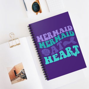 Op de afbeelding: Paarse spiraalnotitieblok met de tekst "Mermaid Mermaid At Heart" in een retro-lettertype met een schelp en een zeemeerminstaart.