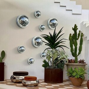 May include: A collection of silver disco ball wall decor in various sizes, mounted on a light-colored wall. The scene includes potted plants, including cacti and succulents, and two decorative ottomans with brown and orange tops. The image is labeled "Set 3".