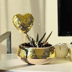 May include: A decorative succulent plant in a gold mosaic pot with a pink base. A gold mosaic heart on a stick is placed in the pot. The items are on a white shelf, with a television in the background.