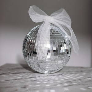 Può includere: Una palla da discoteca argentata con un fiocco di tulle bianco. La palla è ricoperta da piccoli specchi quadrati riflettenti. Il fiocco è legato sulla parte superiore della palla. Lo sfondo è grigio chiaro.