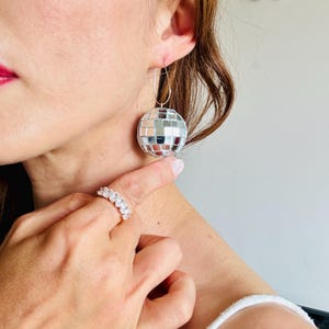 May include: Silver disco ball earrings with a hoop closure. The earrings are round and covered in small, reflective squares. A silver ring with multiple clear stones is also visible.