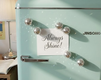May include: A mint green refrigerator with a silver handle and the brand name JINSONG printed on the front. A white square note with the black cursive text Always Shine! is attached to the door by six small, reflective silver disco ball magnets.