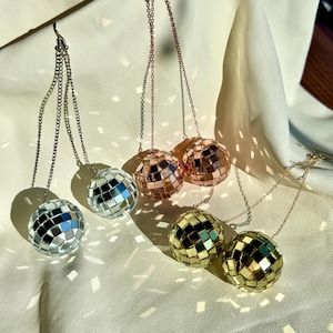 May include: Close-up of four pairs of disco ball earrings. The earrings are silver, rose gold, and gold, each with a chain and a mirrored ball. The earrings are hanging against a white background with shadows.