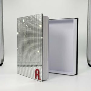 Op de afbeelding: Een zilveren spiegelboek met een rode letter "A" op de kaft, naast een open zwarte en witte doos. De kaft van het boek is gemaakt van kleine, reflecterende vierkantjes. Het boek ligt op een wit oppervlak.