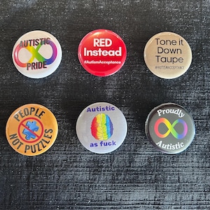 May include: A collection of six round buttons with various designs and text related to autism awareness and pride. The buttons feature phrases like "Autistic Pride," "Red Instead," and "People Not Puzzles," along with the infinity symbol.