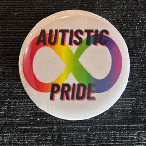 May include: A round button with the words "AUTISTIC PRIDE" in black text. The design features a rainbow-colored infinity symbol. The button has a white background.