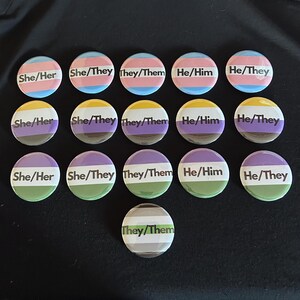 May include: A collection of sixteen circular pin-back buttons arranged in rows. Each button features a horizontal striped flag background in various color combinations with black text displaying pronouns including She/Her, She/They, They/Them, He/Him, and He/They.
