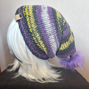 May include: A handmade knit beanie in a striped pattern of yellow, purple, gray, and white. The hat has a purple faux fur pom-pom and a small tag attached. This winter hat is a cozy accessory.