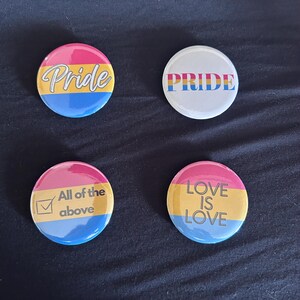 May include: Four circular pin-back buttons featuring pansexual pride flag colors of pink, yellow, and blue. One button says Pride in white script, one says PRIDE in block letters, one says All of the above with a checkbox, and one says LOVE IS LOVE.