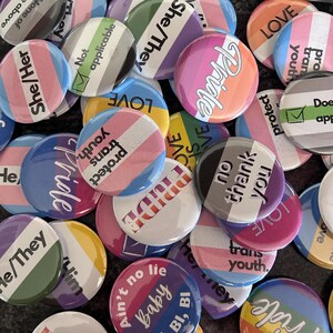 May include: A collection of round pin-back buttons featuring various pride flag color schemes and text including She/Her, He/They, Pride, Love, Protect trans youth, No thank you, Not applicable, and Ain't no lie baby bi, bi. The buttons are arranged in a pile.