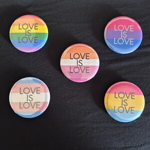 May include: Five circular pin-back buttons featuring various pride flag color schemes. Each button displays the text LOVE IS LOVE in black capital letters. The designs include a rainbow flag, a pink and blue flag, a lesbian flag, a trans flag, and a pan flag.
