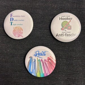 May include: Three circular pinback buttons with white backgrounds. One features text FDT with a yarn ball illustration. Another displays the text Part-Time Hooker Full-Time Anti-fascist with a yarn and hook graphic. The third shows colorful crochet hooks and the word Resist.