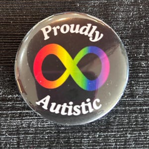 May include: A round button with the words "Proudly Autistic" in white text. The button features a colorful infinity symbol in red, orange, yellow, green, blue, and purple on a black background.