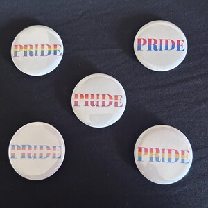 May include: Five circular white pin-back buttons, each featuring the word PRIDE in capital letters. The text on each button displays a different horizontal color stripe pattern, including rainbow, blue and red, orange and red, blue and pink, and yellow, blue, and pink.