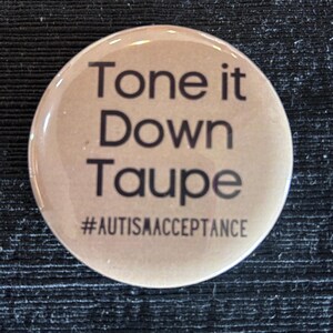 May include: A round, taupe-colored button with the text "Tone it Down Taupe" in black, along with the hashtag "#AUTISMACCEPTANCE". The button has a glossy finish and is likely a pin or badge.