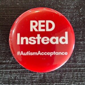 May include: A round, glossy red button with the words "RED Instead" in large white letters. Below, the hashtag "#AutismAcceptance" is printed in white. The button is a symbol of support and awareness.