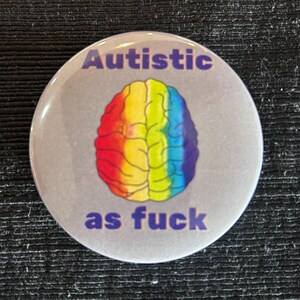 May include: A round button with the text "Autistic as fuck" in purple. The button features a colorful brain graphic in rainbow colors. The background is a light gray.