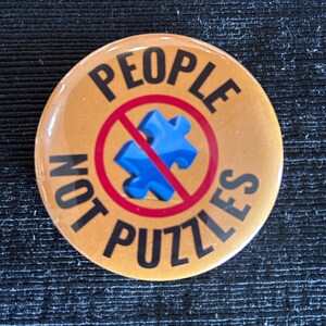 May include: A round button with the words "PEOPLE NOT PUZZLES" in black text on a gold background. A blue puzzle piece is crossed out with a red circle and line.