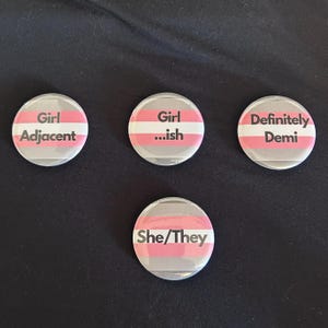 May include: Four circular pin-back buttons featuring a horizontal striped pattern in shades of gray, white, and pink. The buttons display the black text: Girl Adjacent, Girl...ish, Definitely Demi, and She/They, each centered on the striped background.