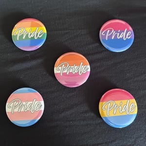 May include: Five circular pin-back buttons featuring various pride flag color patterns. Each button displays the word Pride in a white cursive script font. The designs include rainbow, bisexual, lesbian, transgender, and pansexual flag color schemes.